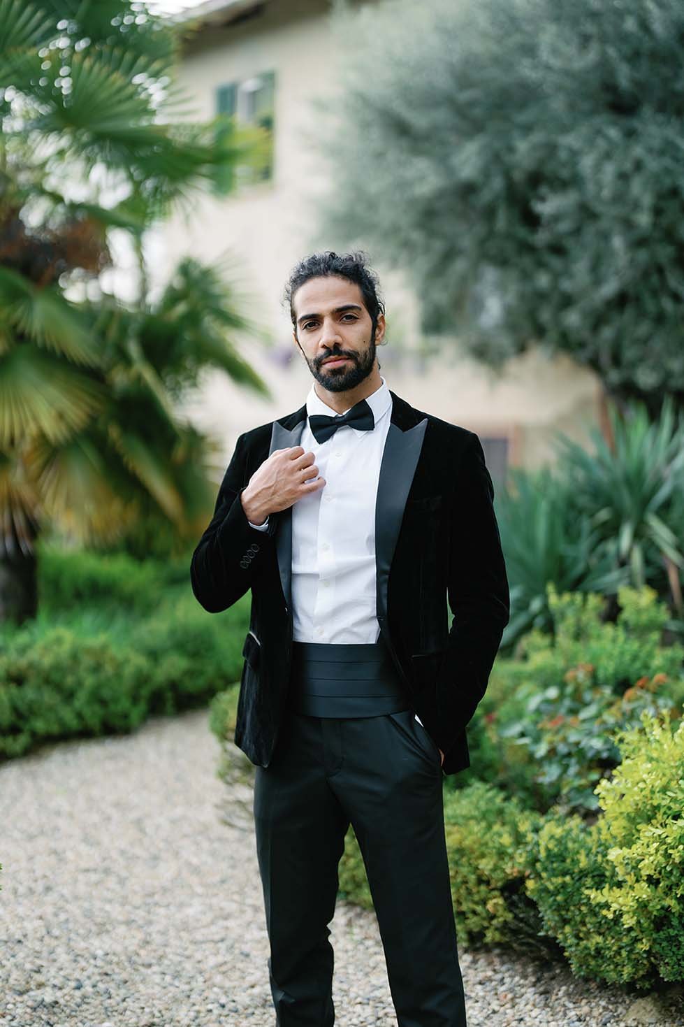 A male model in a black suit with dark long hair adjusts his dark jacket in the background of a castle in the garden