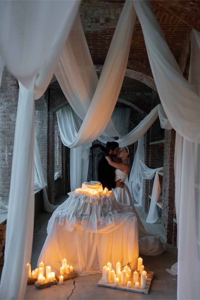 The bride and the groom kissing behind the table with floral decorations in a room with brick walls and white curtains