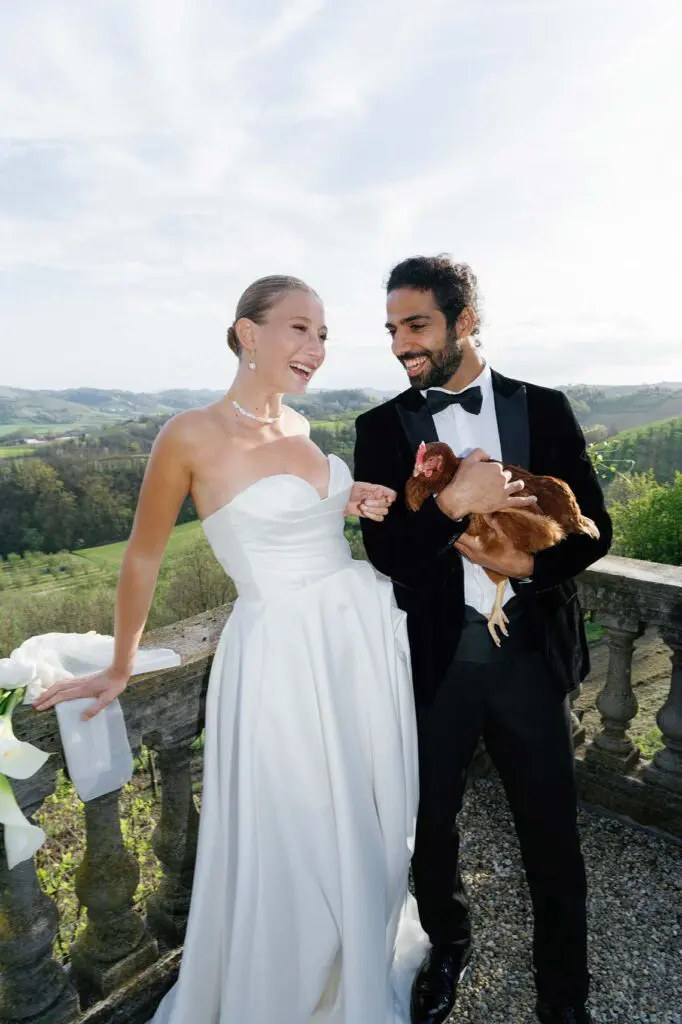 The bride laughs with the groom who is holding a chicken in the background of a beautiful landscape