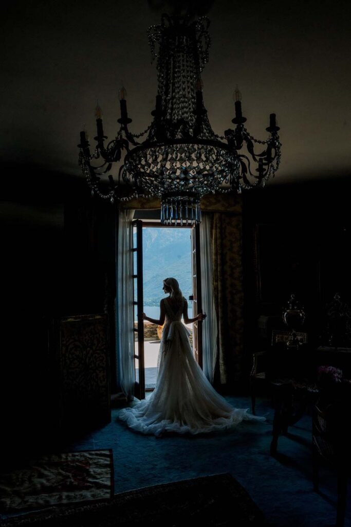 A girl in a gorgeous white dress stands at a doorway with a backdrop of the mountains of Lake Como