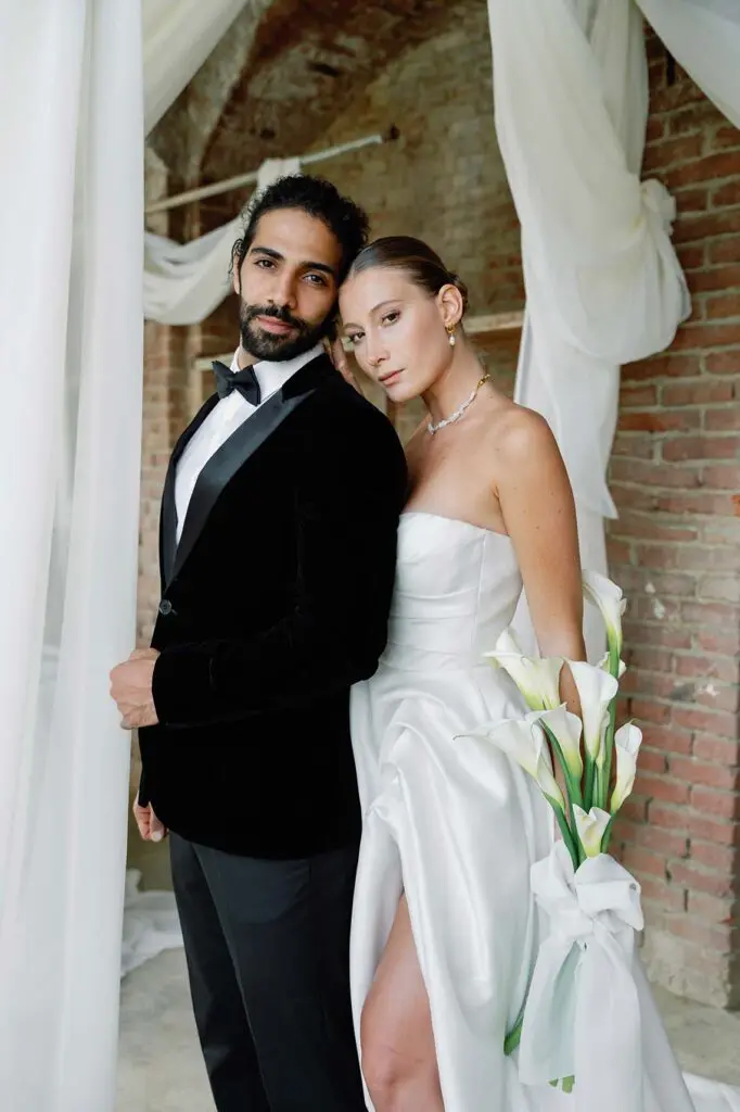 The bride hugs the groom from behind against a backdrop of a brick wall and white curtains