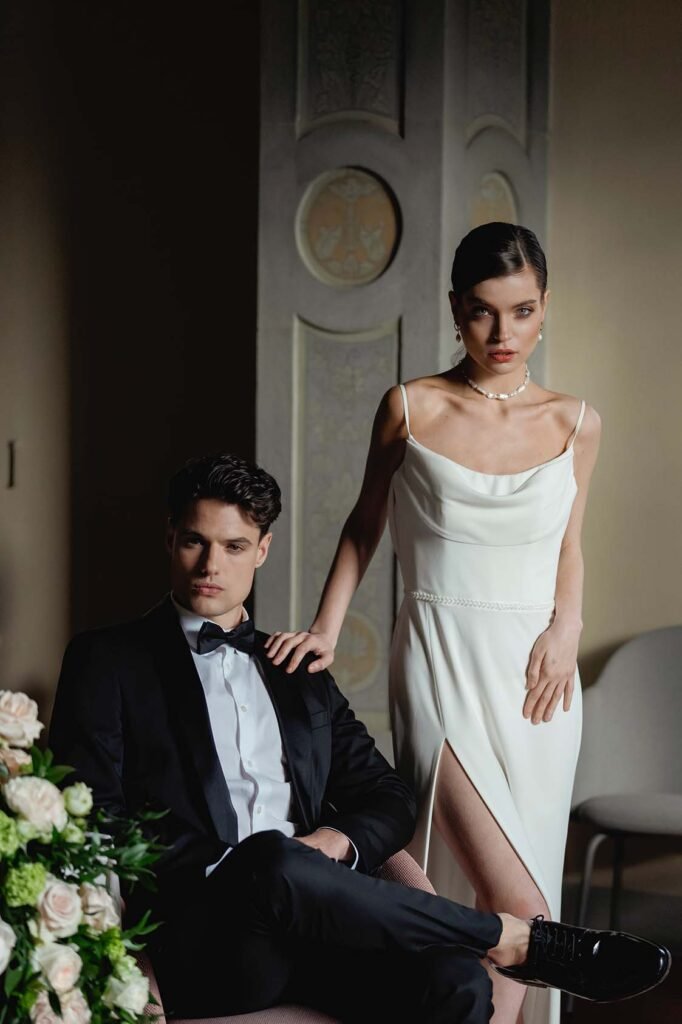 The groom with a serious look is sitting on a chair and his bride is standing next to him with her hand on his shoulder