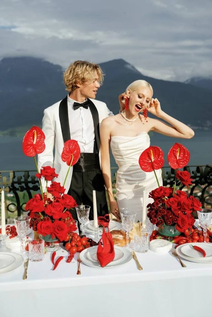 The bride and groom give the bride an “earring” of red berries near a table of red flowers with the mountains of Lake Como as a backdrop