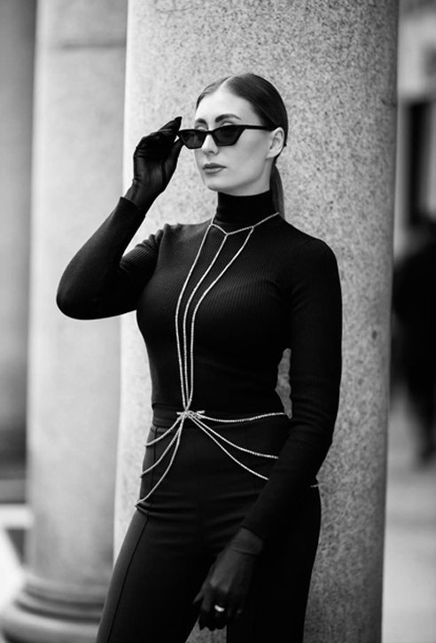 Girl wearing sunglasses with black clothes and dark hair against a stone column in the background.