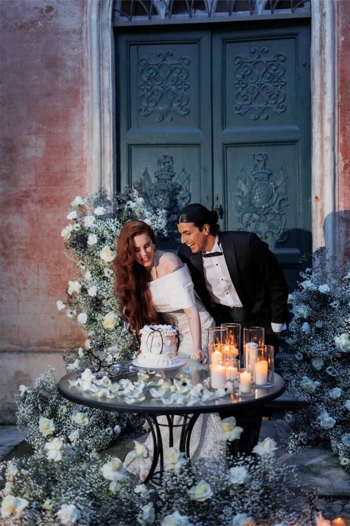 A couple celebrating a wedding, in front of them there is a table with a cake and candles around which there are white decorative flowers