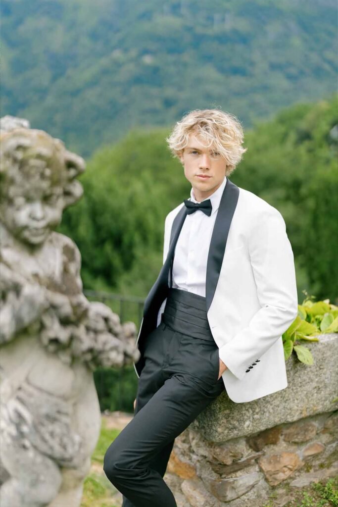 A guy in a white suit with a black bowtie sits on a rock against a background of a statue and trees