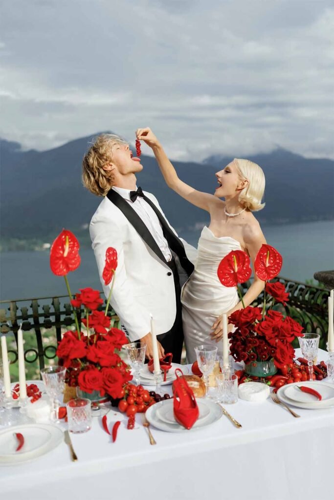 A bride with white hair puts pepper in the groom's mouth in front of a table with a red-colored backdrop of Lake Como mountains