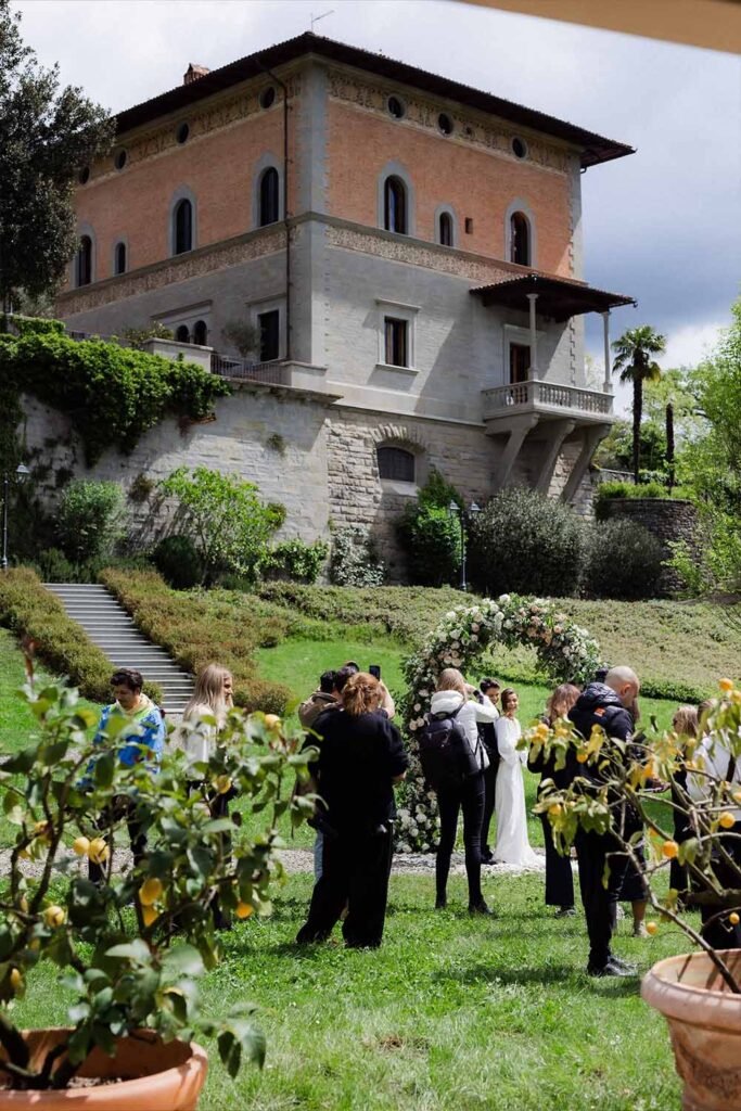 Photographers work together on a photo of a couple standing in front of a villa and an archway of flowers