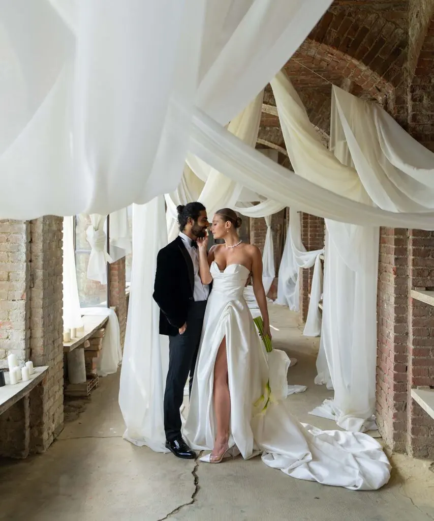 The bride and groom stand in an embrace in a room with brick walls and white curtains and the bride reaches out to kiss the groom