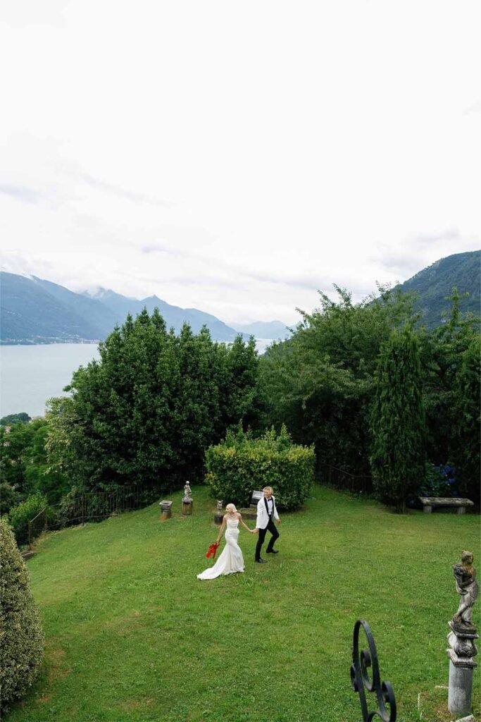 A couple walks holding hands in the garden of a villa against the backdrop of Lake Como
