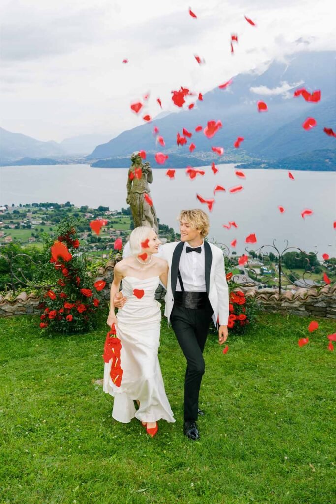 A couple in an embrace walk up a grassy slope with rose petals flying around them