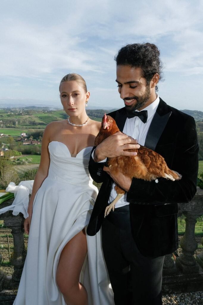 The groom contentedly holds a hen in his arms next to his bride in the background of the landscape