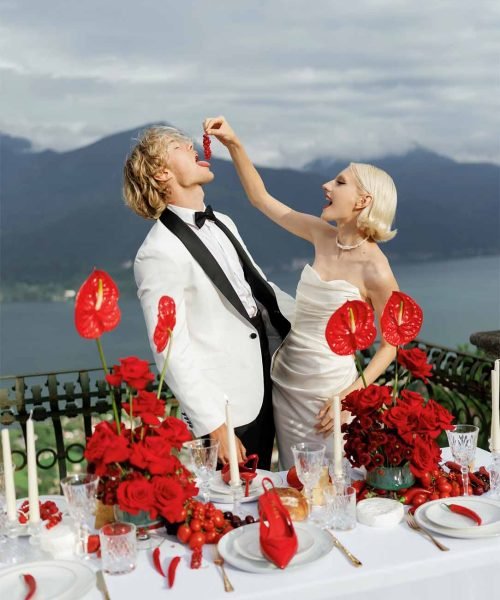 A bride with white hair puts pepper in the groom's mouth in front of a table with a red-colored backdrop of Lake Como mountains