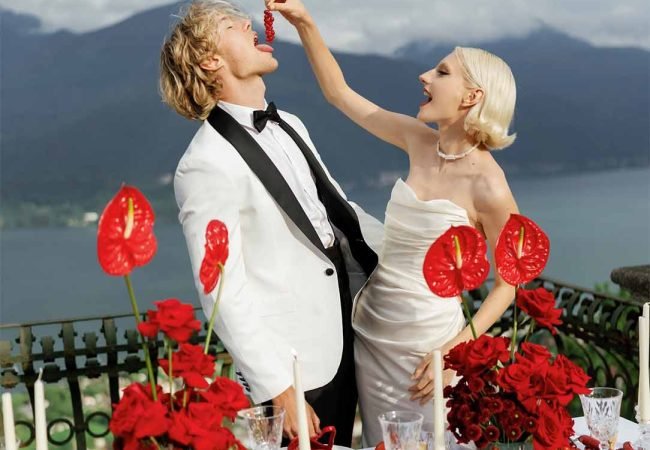A bride with white hair puts pepper in the groom's mouth in front of a table with a red-colored backdrop of Lake Como mountains