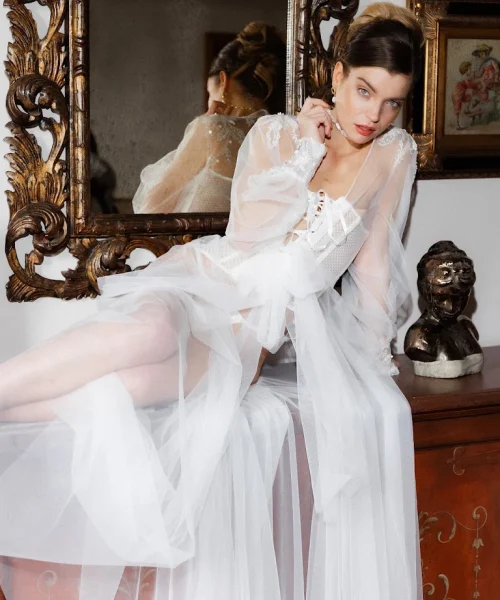 A girl in a gorgeous white dress poses lying on a shelf, and behind her back hangs a mirror in a fancy frame