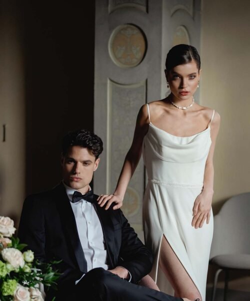 The groom with a serious look is sitting on a chair and his bride is standing next to him with her hand on his shoulder