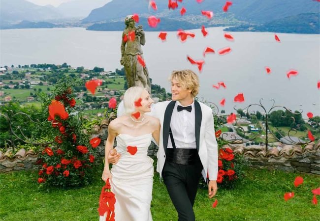 A couple in an embrace walk up a grassy slope with rose petals flying around them
