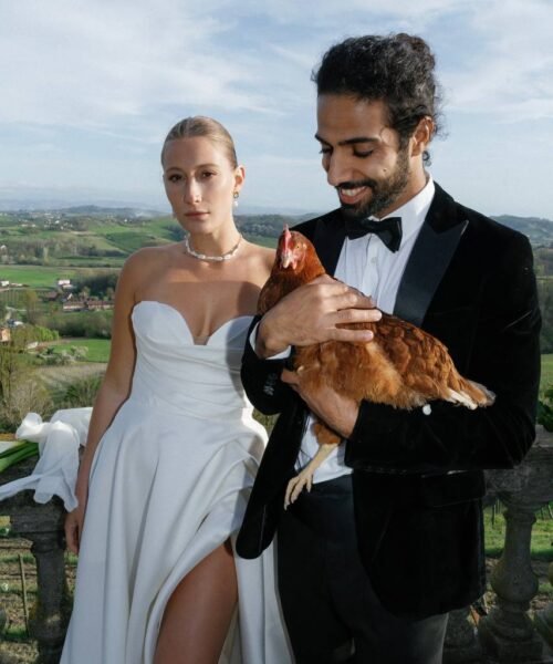 The groom contentedly holds a hen in his arms next to his bride in the background of the landscape